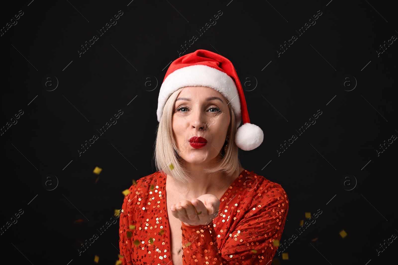 Photo of Woman with perfect makeup in Santa hat blowing confetti on black background. Christmas celebration