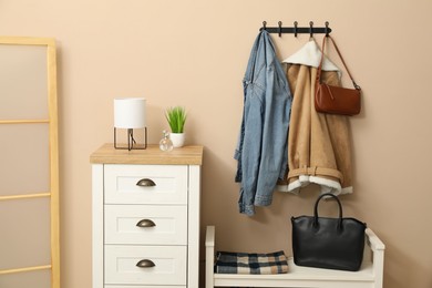 Photo of Coat rack with clothes, mirror, shoe storage bench and chest of drawers in hallway. Interior design