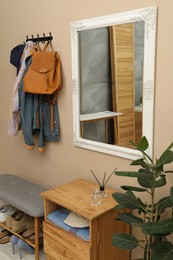 Coat rack with clothes, mirror, houseplant and shoe storage bench in hallway. Interior design