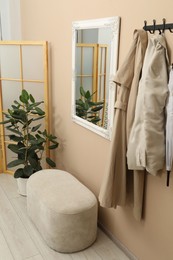 Photo of Coat rack with clothes, pouffe, houseplant and mirror in hallway. Interior design