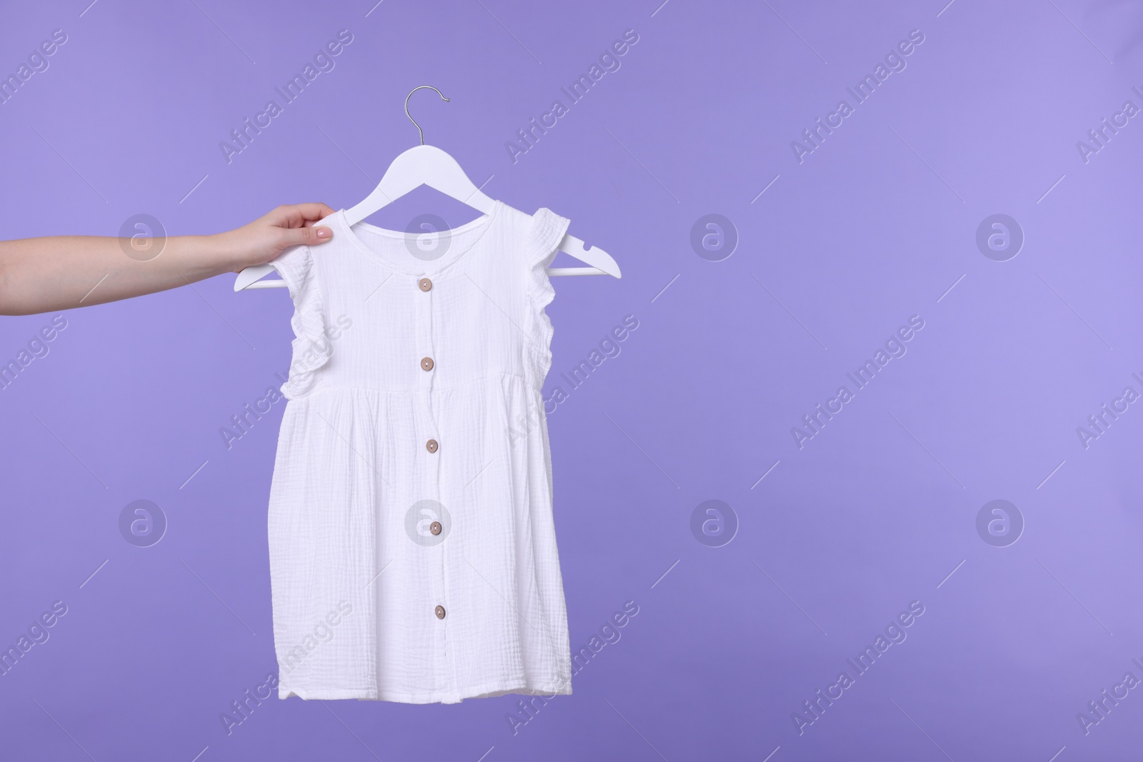 Photo of Woman holding hanger with white baby dress on light purple background, closeup. Space for text