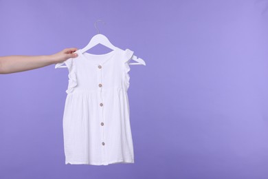 Photo of Woman holding hanger with white baby dress on light purple background, closeup. Space for text