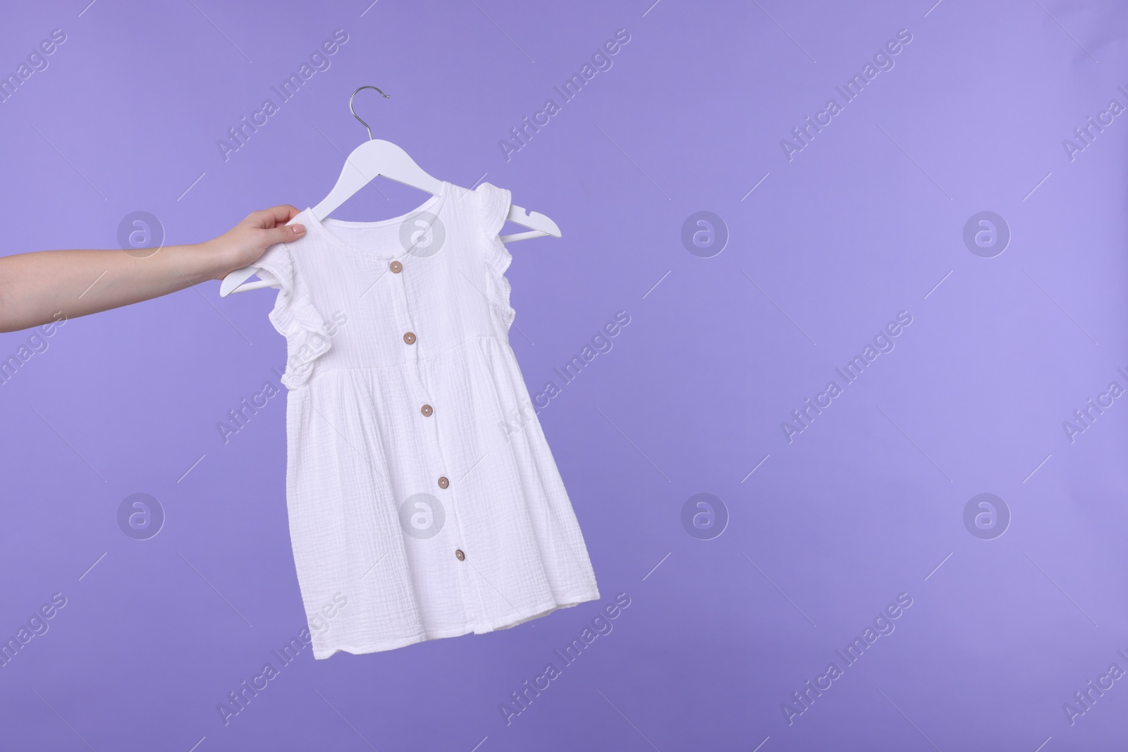 Photo of Woman holding hanger with white baby dress on light purple background, closeup. Space for text