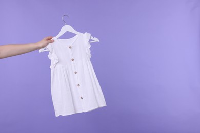 Photo of Woman holding hanger with white baby dress on light purple background, closeup. Space for text