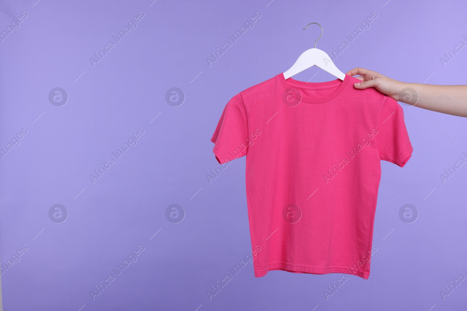 Photo of Woman holding hanger with pink t-shirt on light purple background, closeup. Space for text