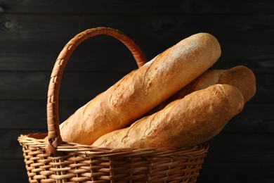 Photo of Fresh baguettes in wicker basket against black wooden background, closeup