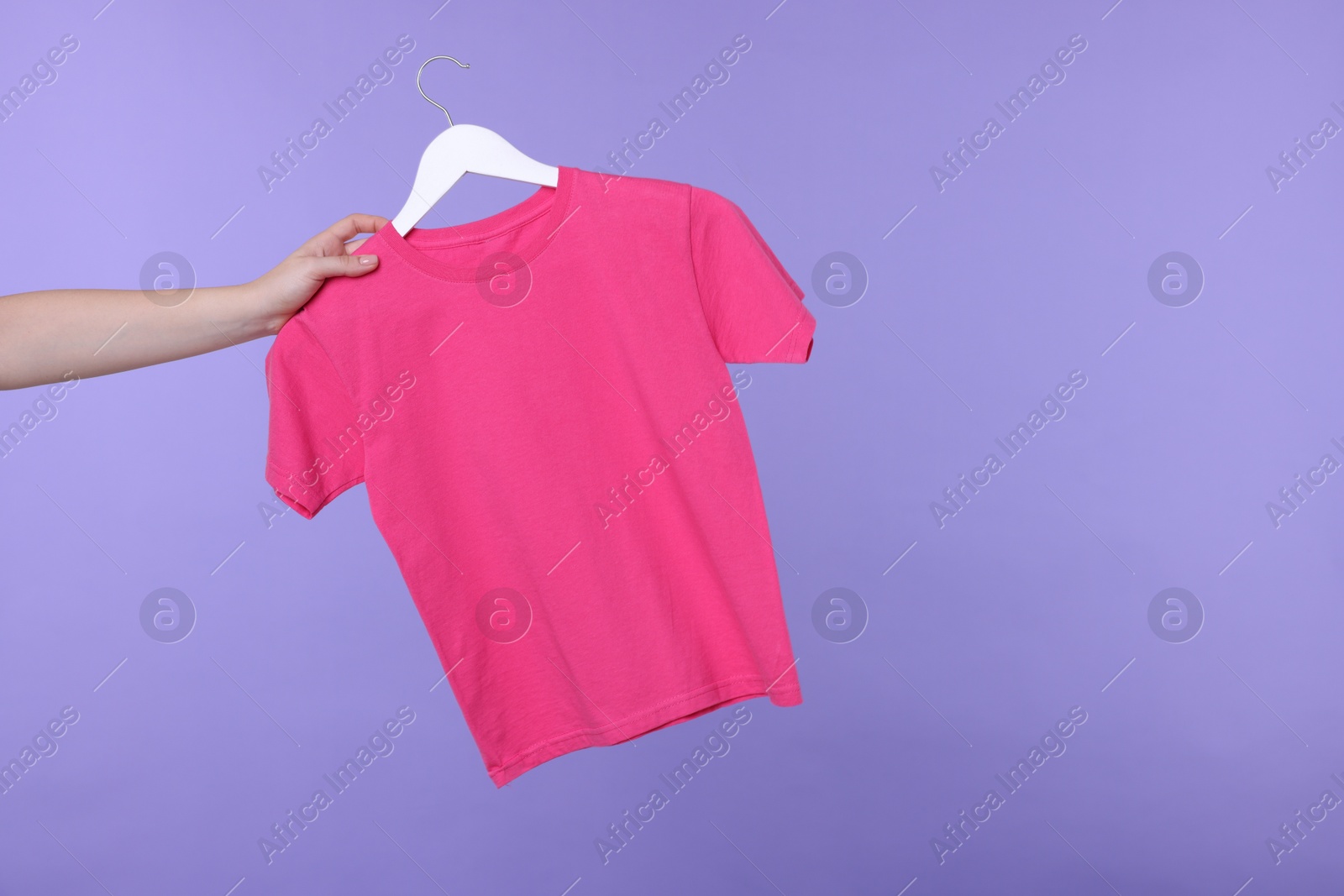 Photo of Woman holding hanger with pink t-shirt on light purple background, closeup. Space for text