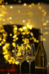 Photo of Glasses and bottle of white wine against blurred Christmas lights, closeup