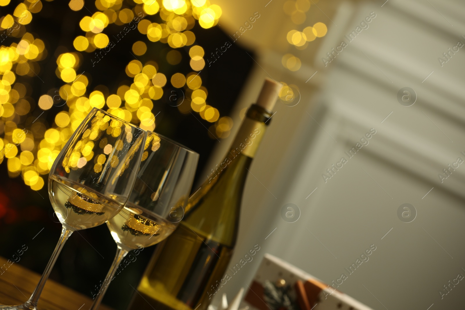 Photo of Glasses and bottle of white wine against blurred Christmas lights, closeup