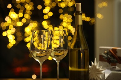 Photo of Glasses and bottle of white wine against blurred Christmas lights, closeup