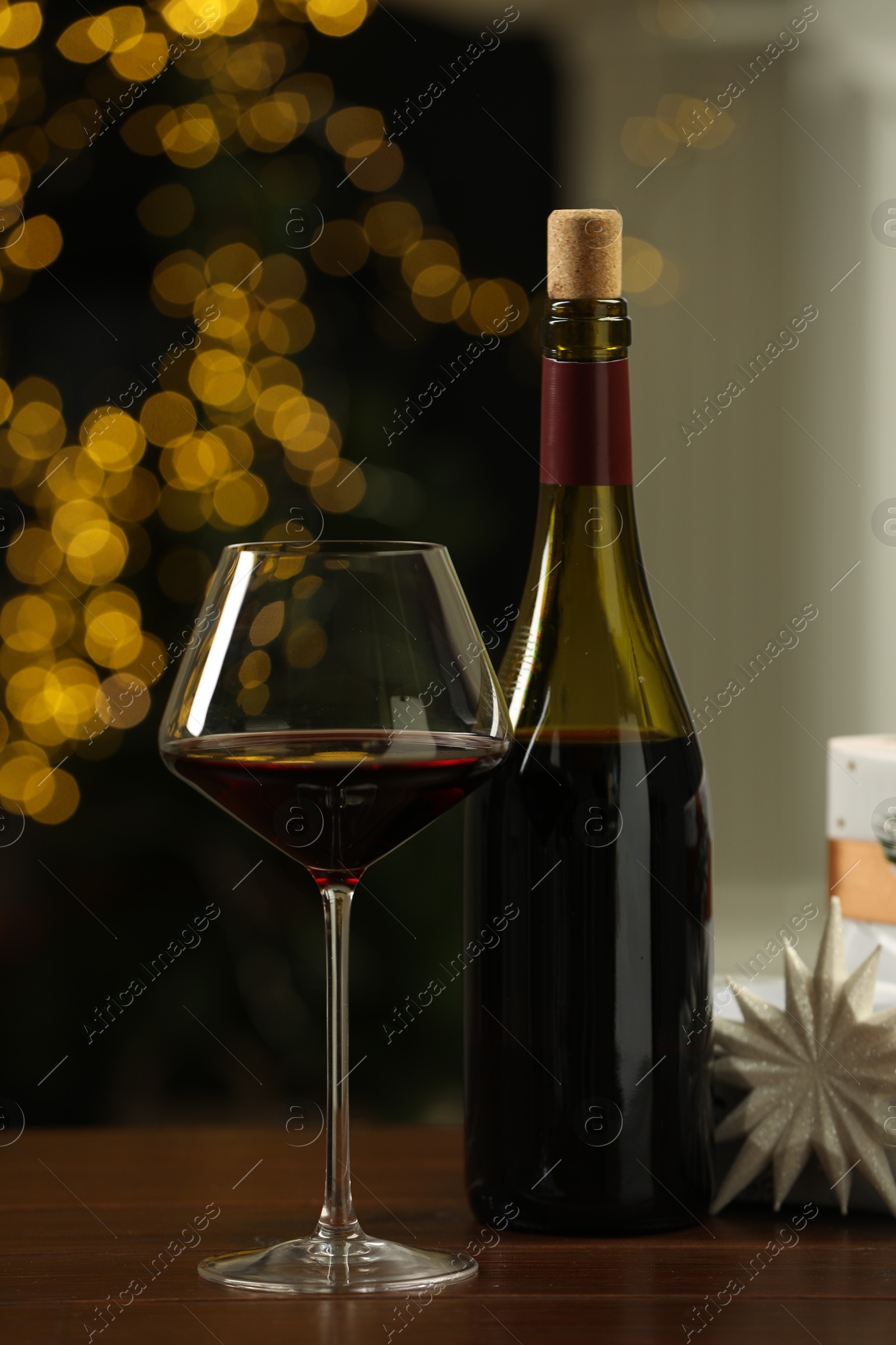 Photo of Glass and bottle of red wine on wooden table against blurred Christmas lights