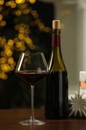 Photo of Glass and bottle of red wine on wooden table against blurred Christmas lights