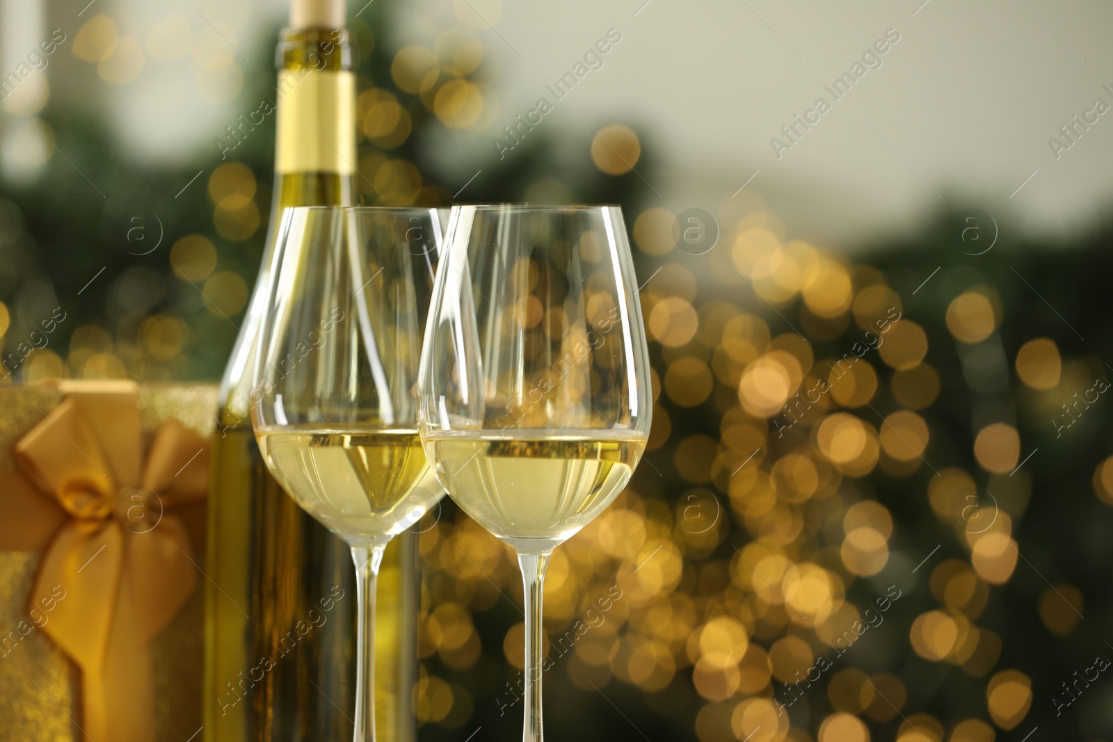 Photo of Glasses and bottle of white wine against blurred Christmas lights, closeup. Space for text