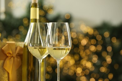 Photo of Glasses and bottle of white wine against blurred Christmas lights, closeup. Space for text