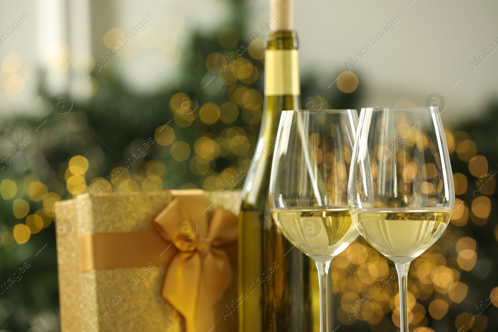 Photo of Glasses and bottle of white wine against blurred Christmas lights, closeup