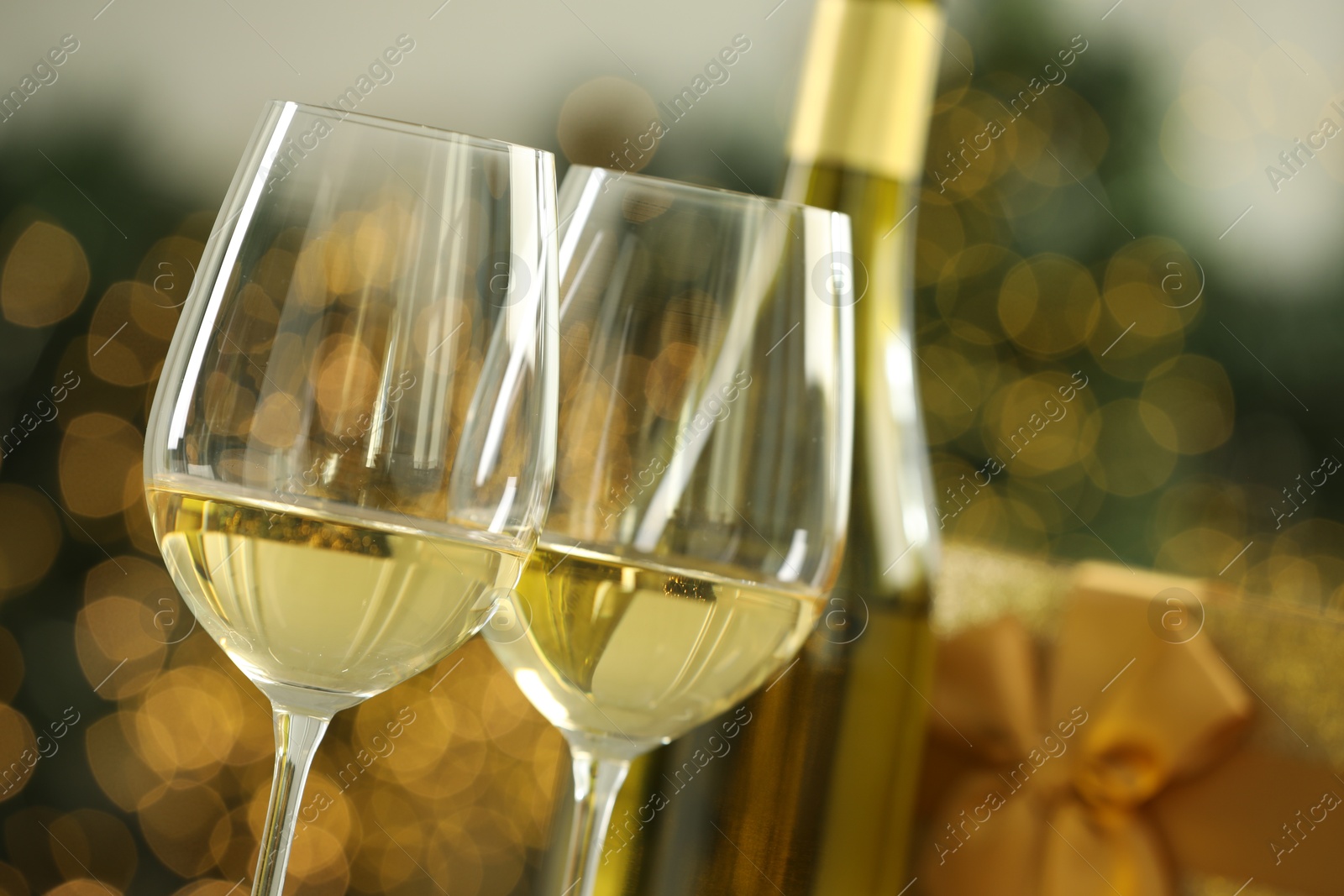 Photo of Glasses and bottle of white wine against blurred Christmas lights, closeup