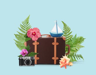 Image of Leather travel suitcase, toy sailboat, seashells, sea star, camera, tropical leaves and flowers on light blue background