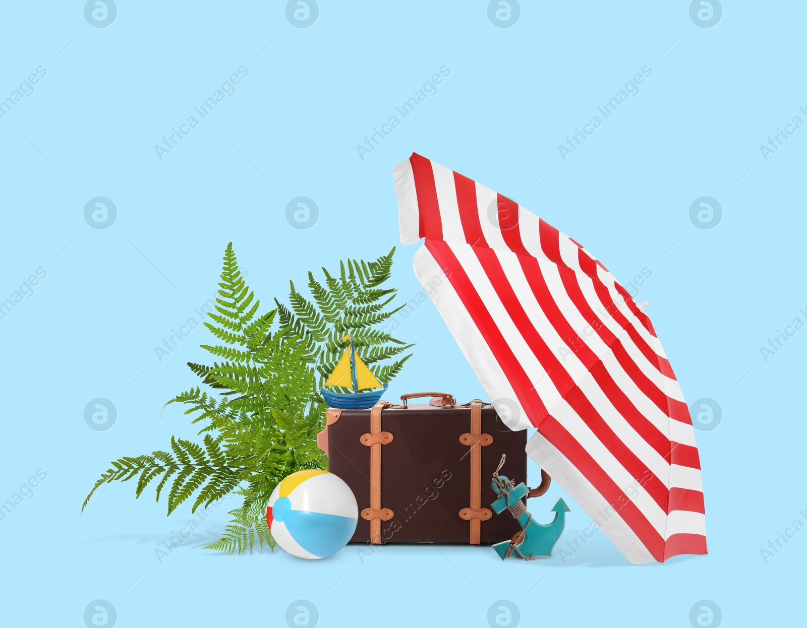 Image of Leather travel suitcase, tropical leaves, toy sailboat and anchor, beach umbrella and ball on light blue background