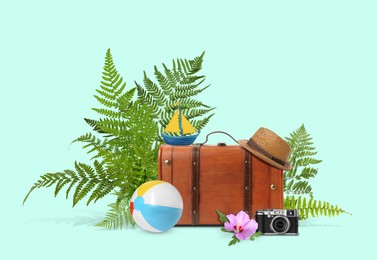 Image of Brown travel suitcase, hat, camera, toy sailboat, beach ball, tropical leaves and flower on light blue background