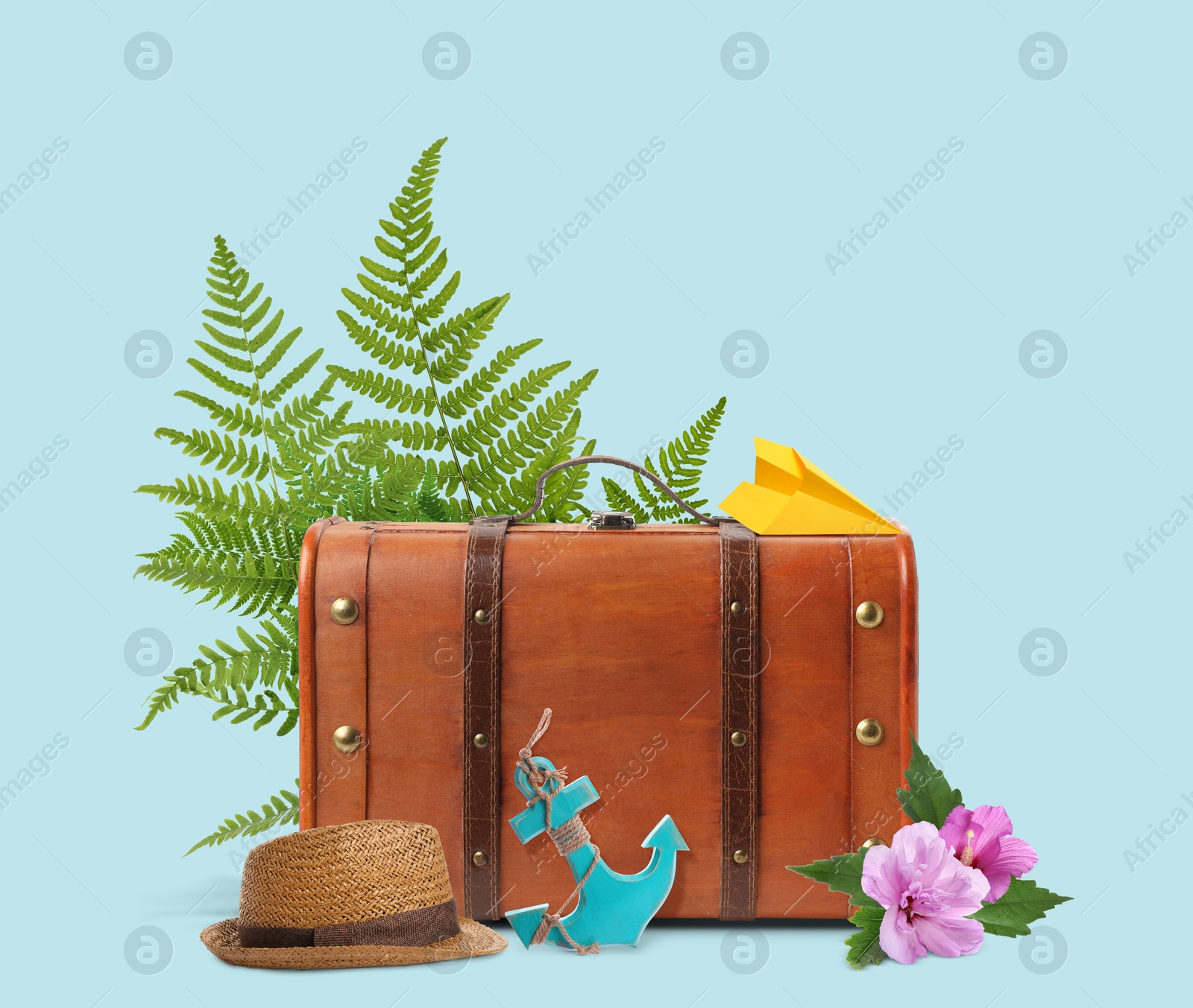 Image of Brown travel suitcase, paper plane, toy anchor, hat, tropical leaves and flowers on light blue background