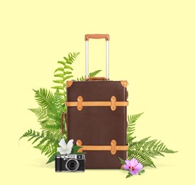 Image of Brown travel suitcase, camera, tropical leaves and flowers on light yellow background