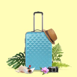 Light blue travel suitcase, hat, camera, seashell, paper boat, tropical leaves and flower on light yellow background