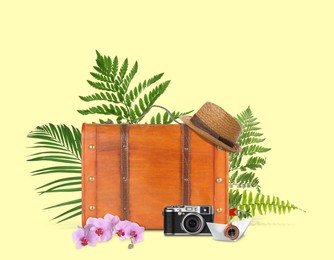 Image of Brown travel suitcase, camera, paper boat, hat, tropical leaves and flowers on light yellow background