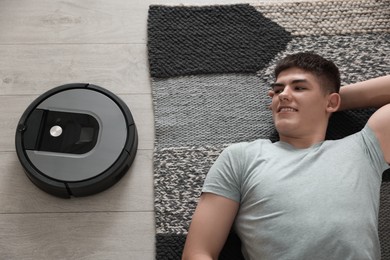 Photo of Robotic vacuum cleaner cleaning floor while smiling man relaxing on carpet, top view