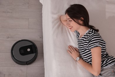 Photo of Smiling young woman resting on bed while robotic vacuum cleaner vacuuming floor at home, top view