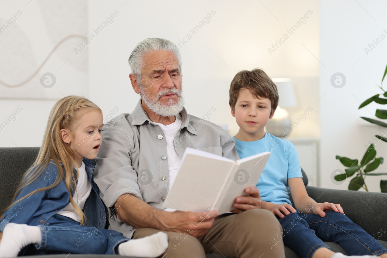 Photo of Grandpa and his grandkids reading book together on sofa at home