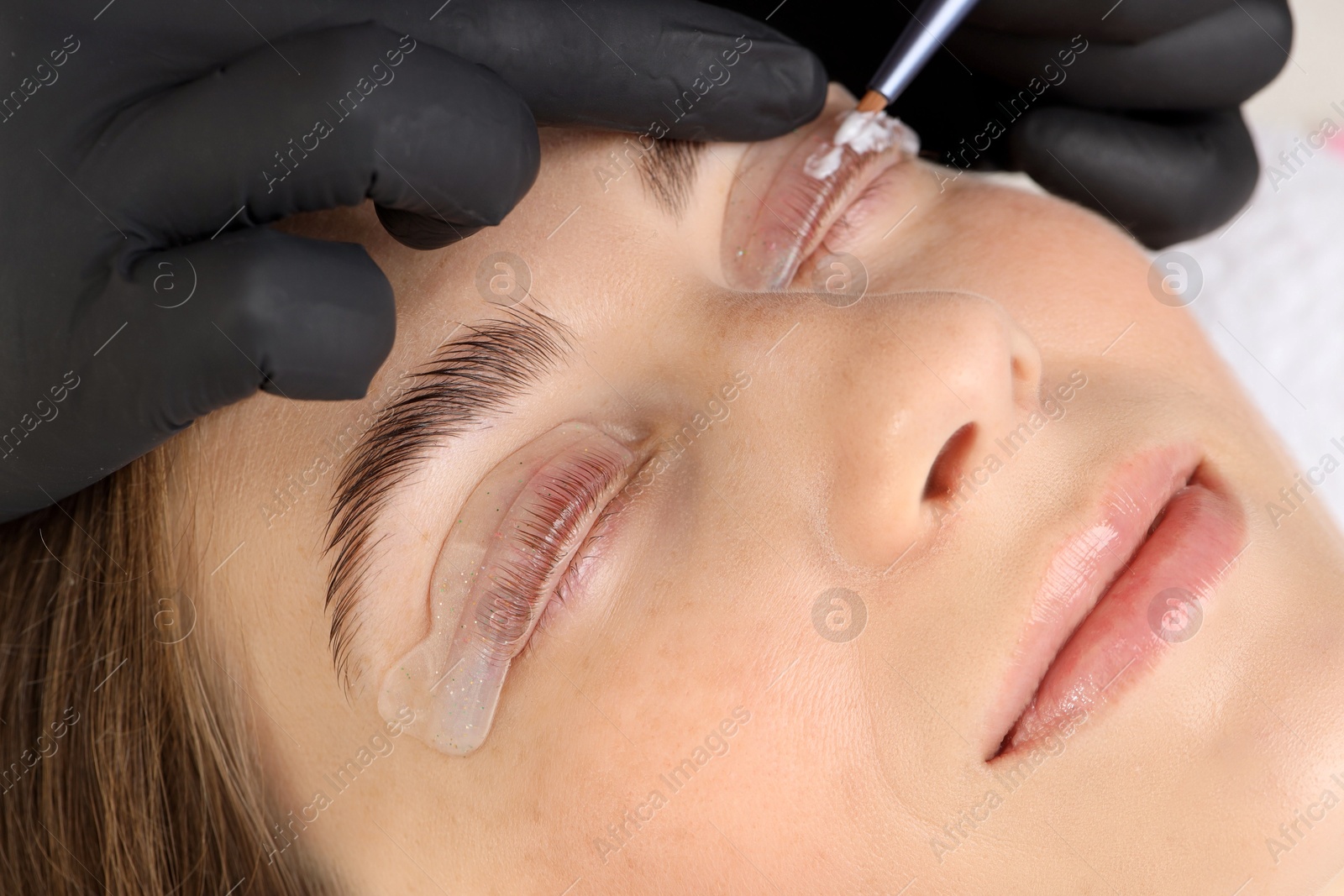 Photo of Eyelash lamination procedure. Esthetician applying lotion on woman's lashes in beauty salon, closeup