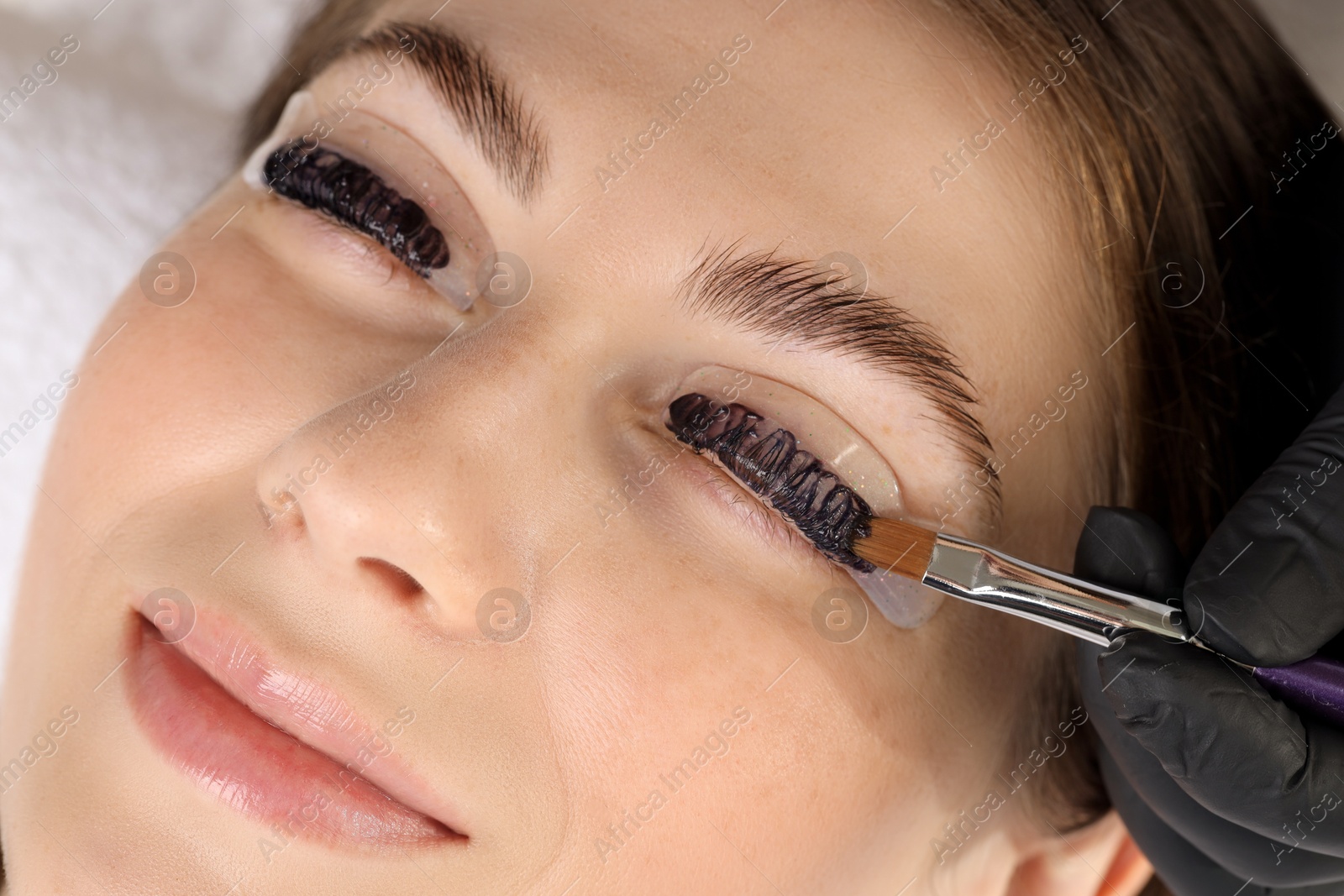 Photo of Eyelash lamination procedure. Esthetician applying dye on woman's eyelashes in beauty salon, closeup