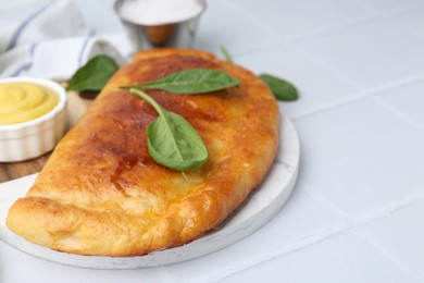 Photo of Tasty calzone with basil and sauce on white tiled table, closeup. Space for text