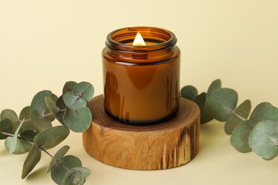 Photo of Beautiful burning candle and eucalyptus branches on pale yellow background, closeup