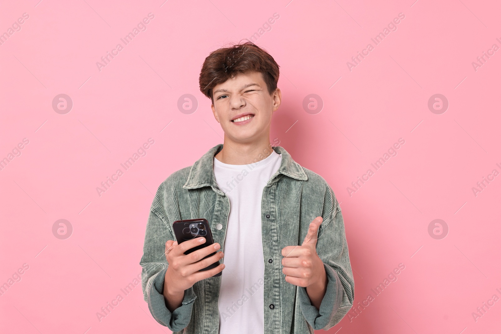 Photo of Happy teenage boy with smartphone showing thumbs up on pink background