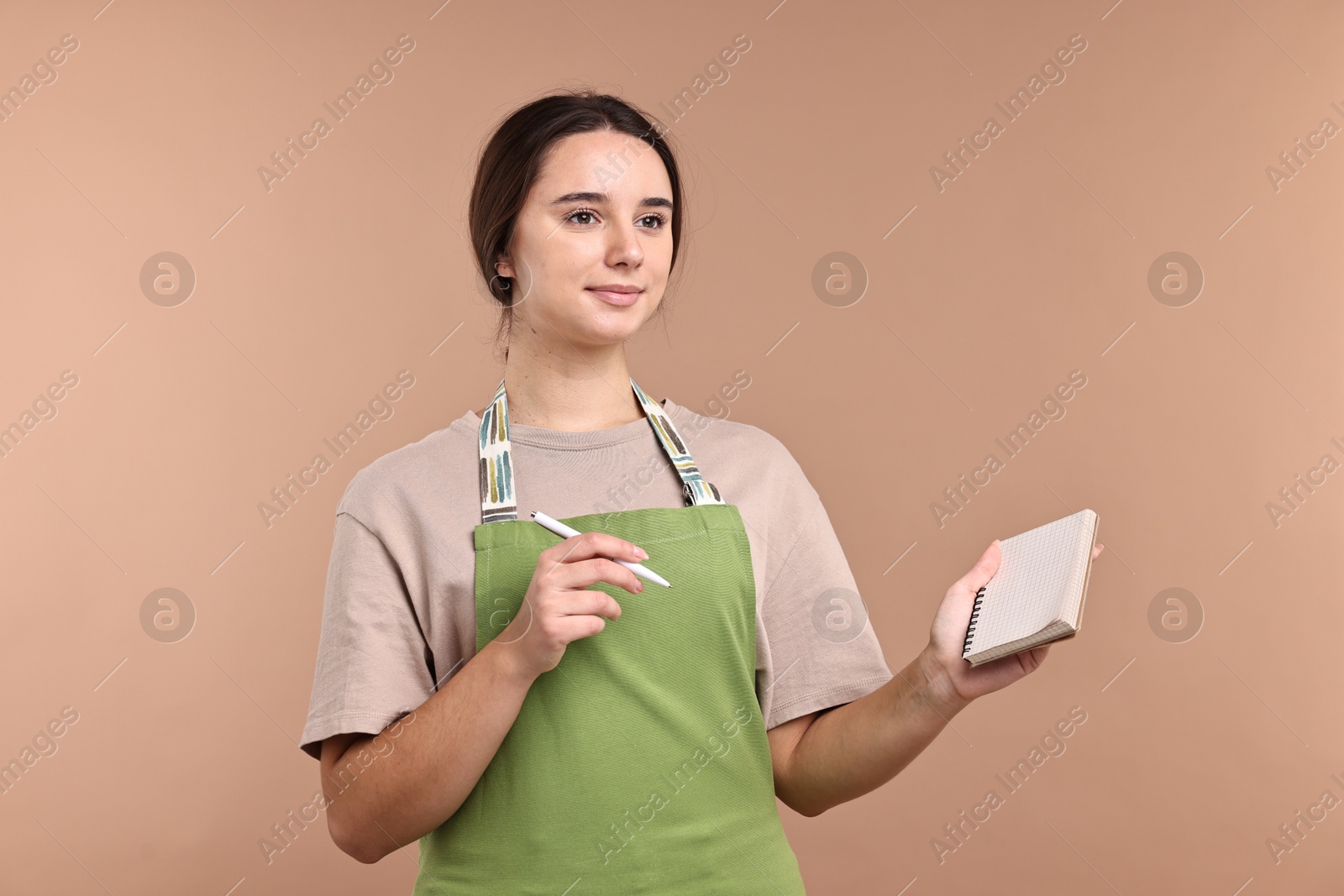 Photo of Girl in apron with pen and notebook on pale brown background. Work for teenagers