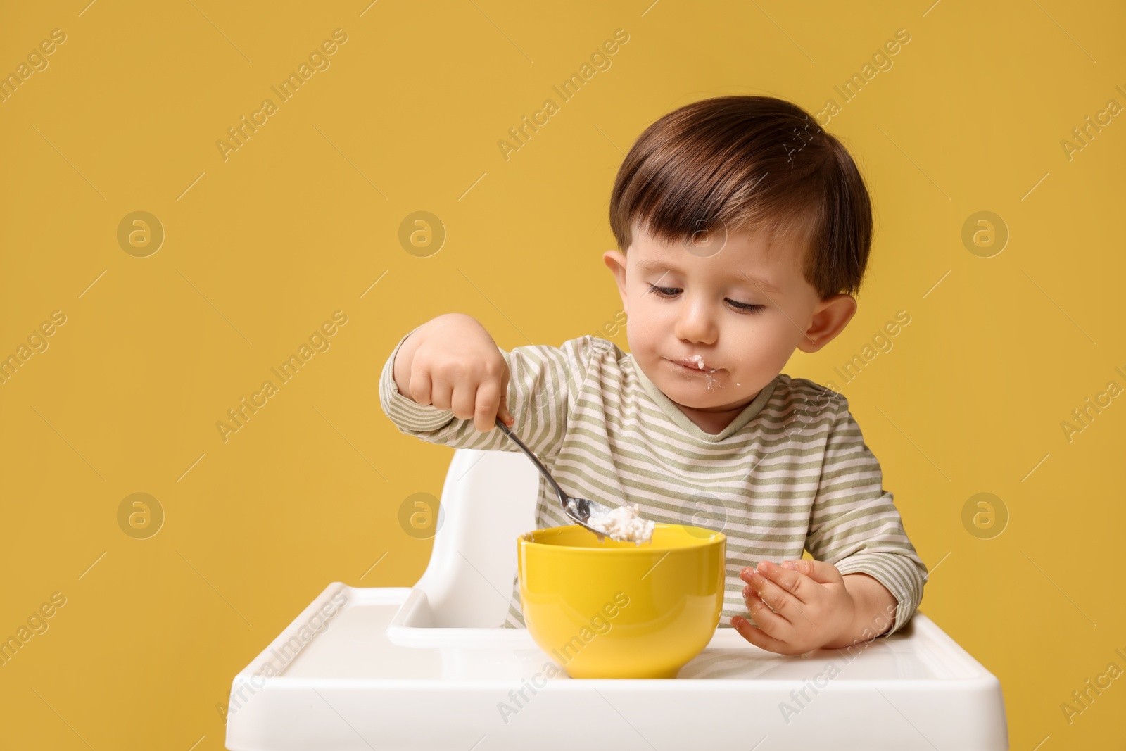 Photo of Cute little kid eating healthy baby food from bowl in high chair on yellow background