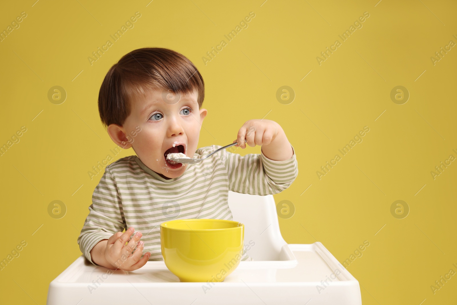 Photo of Cute little kid eating healthy baby food from bowl in high chair on yellow background