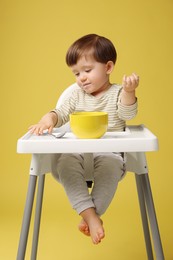 Photo of Cute little kid eating healthy baby food from bowl in high chair on yellow background