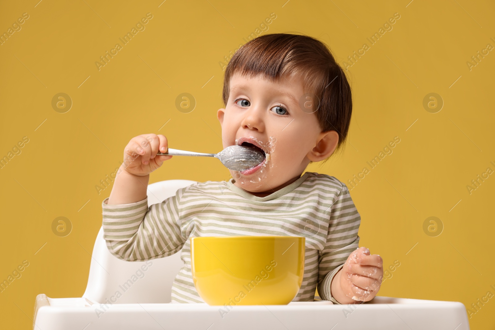 Photo of Cute little kid eating healthy baby food from bowl in high chair on yellow background