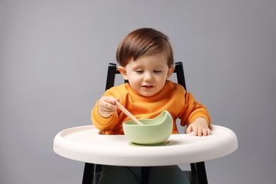 Cute little kid eating healthy baby food from bowl in high chair on light grey background