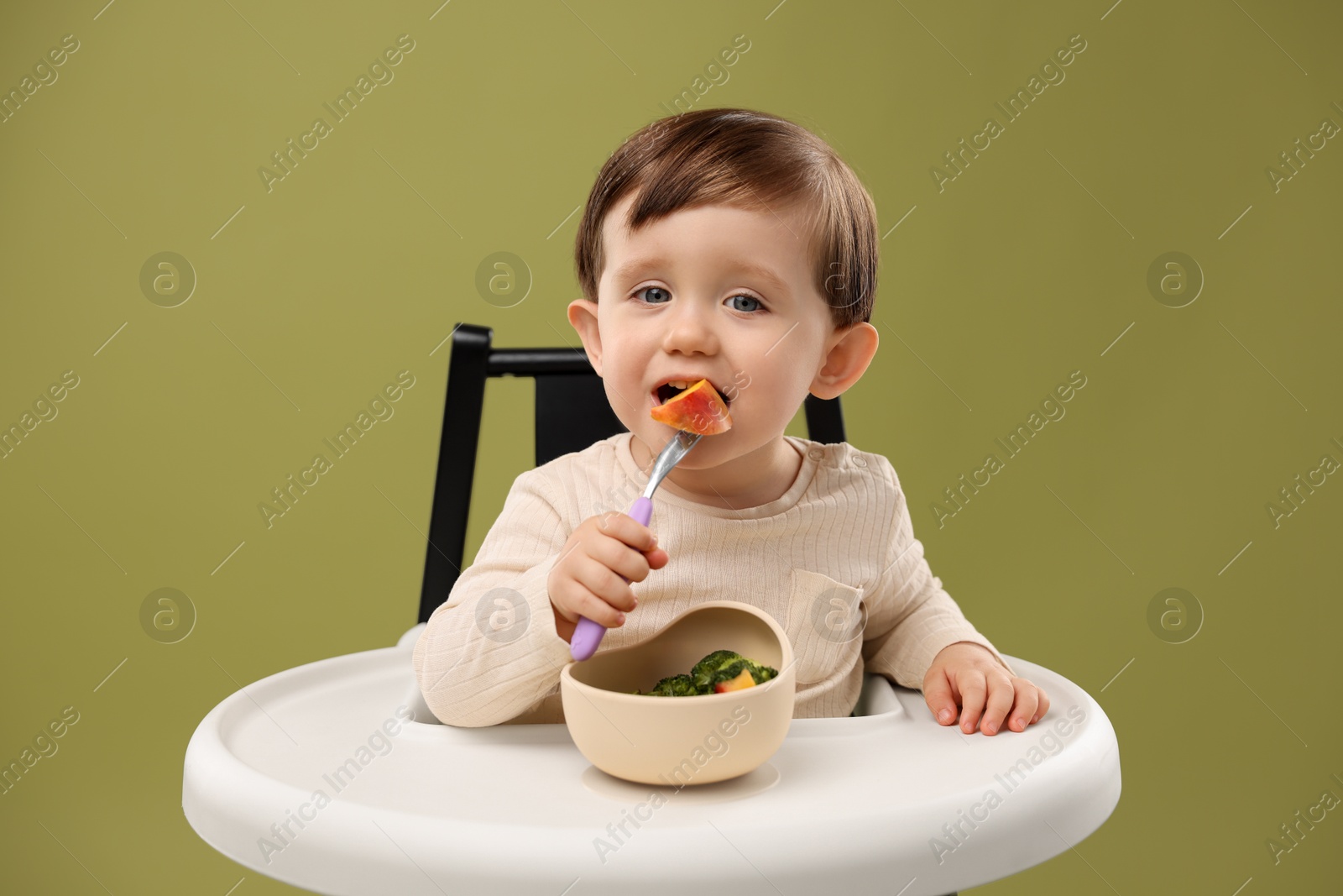 Photo of Cute little baby eating healthy food from bowl in high chair on olive background