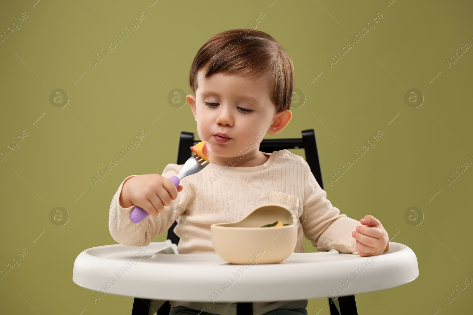 Photo of Cute little baby eating healthy food from bowl in high chair on olive background