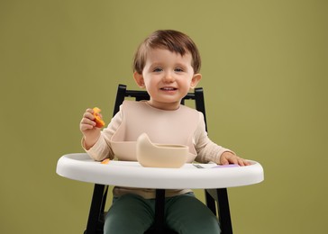 Photo of Cute little baby eating healthy food from bowl in high chair on olive background