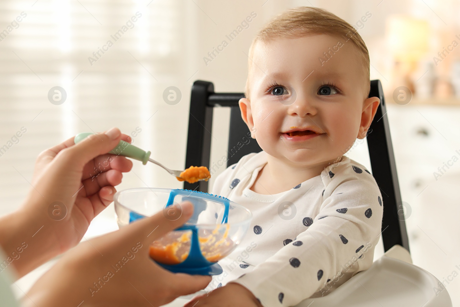 Photo of Mother feeding her cute little baby in high chair at home, closeup