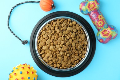 Photo of Dry pet food in feeding bowl and toys on light blue background, flat lay