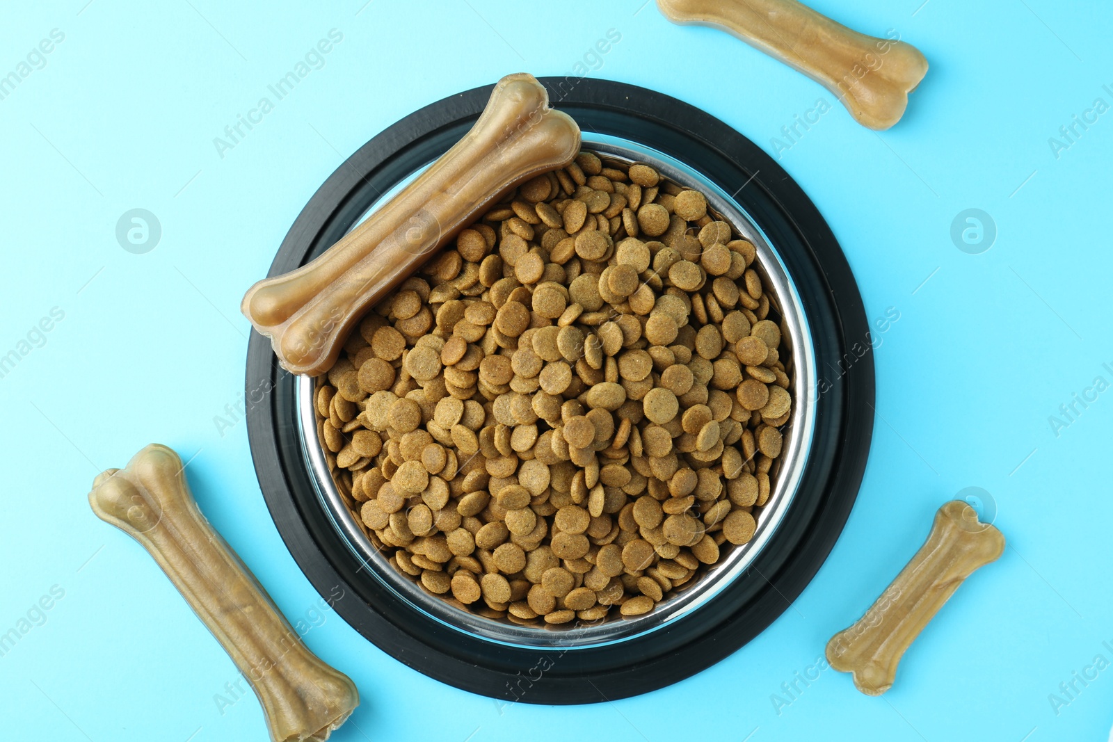 Photo of Dry pet food in feeding bowl and chew bones on light blue background, top view