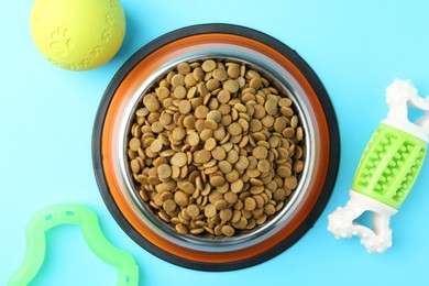 Photo of Dry pet food in feeding bowl and toys on light blue background, flat lay