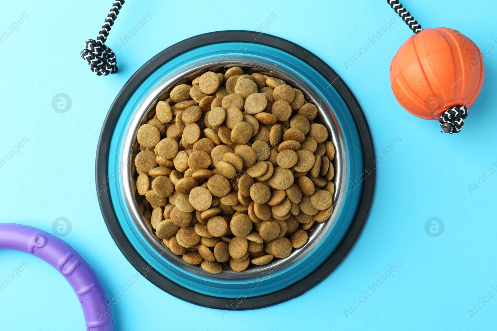 Photo of Dry pet food in feeding bowl and toys on light blue background, flat lay