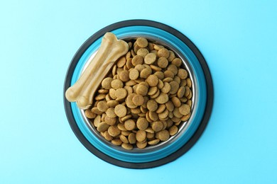 Photo of Dry pet food in feeding bowl and chew bone on light blue background, top view